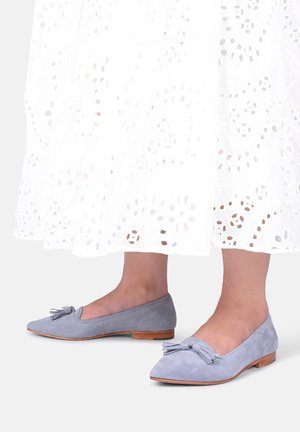 Person iført lyse lilla ruskind flade sko med frynser og en hvid øjlet blonder nederdel, vist fra midt på læggen og ned mod en lys baggrund.