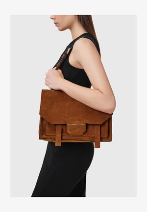 Brown suede tote bag with a folded flap design, accented with gold hardware and hanging straps, held against a black garment.