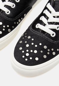 Black sneakers with a textured fur finish. Decorated with metallic studs in various sizes. Features white laces and rubber soles.