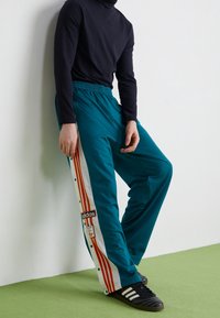 Teal sportbyxor med vita sidsömmar som har tre röda ränder och en Adidas-logotyp; elastisk midja; slätt tyg; bärs med svarta skor.