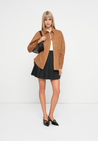 Brown suede button-up shirt over a white top, paired with a black pleated skirt and black pointed-toe heels. Features a small black handbag.