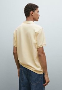 Žluté bavlněné polo tričko s krátkým límcem, umístěné na modelu stojícím bokem, kombinované s tmavě modrými džíny.