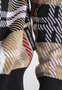 Strikket sweater med en tan-basis og geometriske mønstre i sort, hvid og rød. Sorte ribbede manchetter og et mærketag synligt på siden.