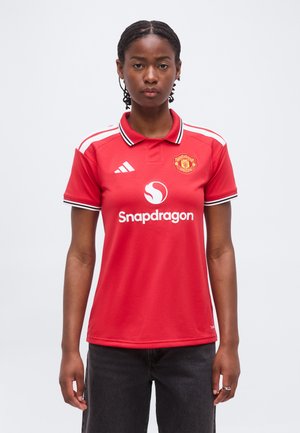 Młoda osoba z warkoczami, ubrana w czerwoną koszulkę piłkarską Manchester United z logo Snapdragon, stoi na jednolitym jasnym tle.