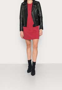 Schwarze Leder-Bikerjacke über einem roten Kleid mit figurbetonter Silhouette. Das Model trägt schwarze Knöchelstiefel. Glatte Textur und minimalistische Details.