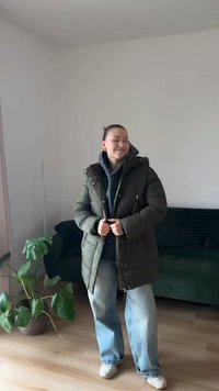 Casaco acolchoado verde-oliva com capuz, fecho de correr frontal e bolsos laterais, usado sobre uma camisola cinza com capuz, combinado com jeans azul claro.