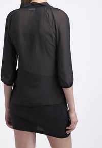 Kvinna i en genomskinlig svart blus med 3/4 ärmar och en figurnära svart kjol, sedd bakifrån mot en enkel ljus bakgrund.