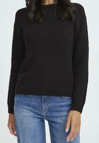 Kvinde iført en sort struktureret strikket sweater og blå jeans, stående mod en lys baggrund med hænderne afslappet langs siden.