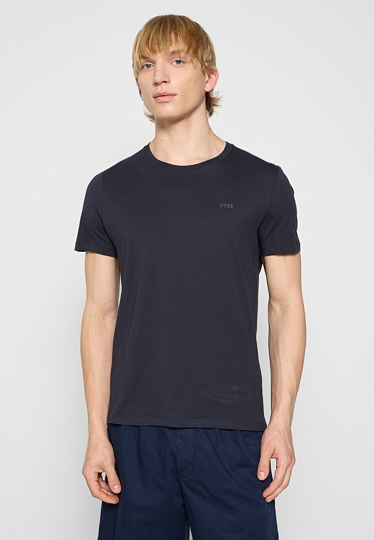 Junger Mann mit blonden Haaren, der ein schlichtes schwarzes BOSS-T-Shirt und eine dunkelblaue Shorts trägt, steht vor einem weißen Hintergrund.