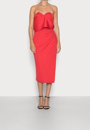 Woman wearing strapless red midi dress with ruffle detail and black ankle-strap high heels, standing against a plain white background.