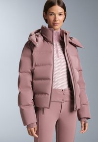 Napuhana ružičasta jakna s visokim ovratnikom, zatvaranjem na patentni zatvarač i rukavima s kopčama uparena s pripijenim topom i hlačama u istom tonu s suptilnim uzorcima.