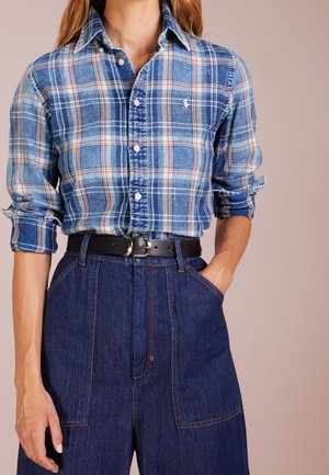 Kvinde iført blå ternet skjorte med knapper og opsmøgede ærmer, mørke jeans med høj talje og sort bælte med sølvspænde, hånd i lommen.