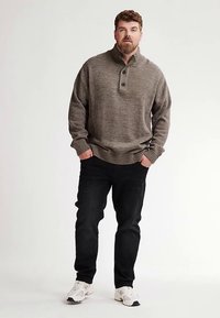 Grauer Strickpullover mit hohem Kragen und drei Knöpfen an der Knopfleiste, kombiniert mit schwarzer Jeans und weißen Turnschuhen. Dicke, gerippte Bündchen und Saum.