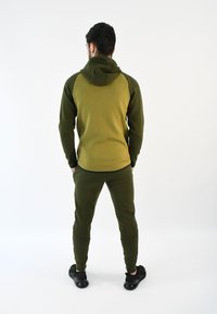 Chándal atlético verde oliva con parte superior con capucha y pantalones ajustados. Presenta puños ceñidos y una textura suave, combinado con zapatillas deportivas negras.