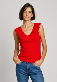 Top rosso senza maniche con scollo a V arricciato, texture a costine e design aderente. Abbinato a jeans blu, mani in tasca.
