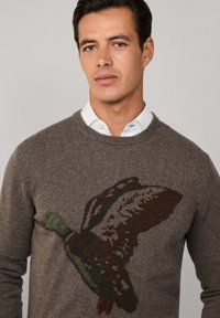Maglione marrone con un design di anatra in volo lavorato a maglia verde e marrone. Stile a girocollo con una texture liscia. Sottile bordo a costine su polsini e orlo.
