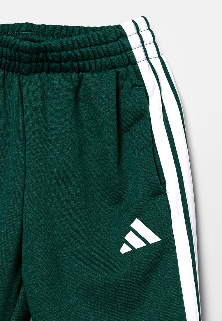 Grüne Baumwoll-Jogginghosen mit einem elastischen Bund, weißen Seitenstreifen, einer Seitentasche und einem weißen Logo am unteren linken Bein.