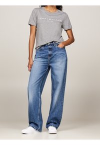 Person i grå Tommy Hilfiger t-shirt, løse blå jeans og hvide sneakers, stående med den ene hånd i lommen foran en ensfarvet baggrund.