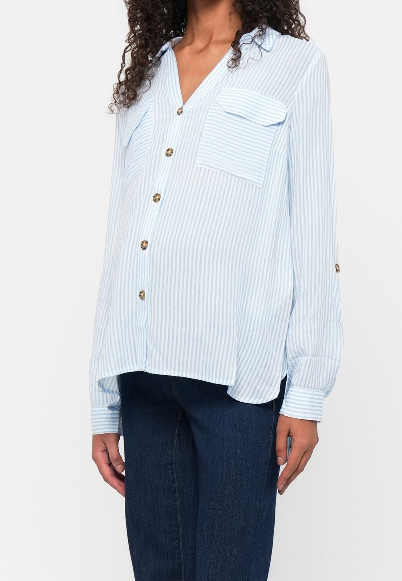Femme portant une chemise à rayures bleu clair et blanc, boutonnée, avec poches sur le devant et manches longues, associée à un jean bleu foncé.