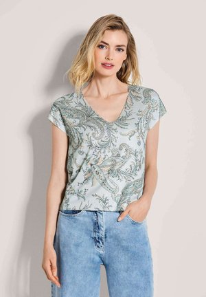 Frau mit blondem Haar, die eine hellblaue Bluse mit floralem Paisley-Muster, V-Ausschnitt und kurzen Ärmeln sowie hellblaue Jeans trägt.