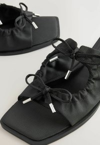 Sandales noires avec une tige texturée noire, présentant une ouverture arrondie, un élastique froncé et des accents métalliques argentés sur les lacets. Semelle plate.