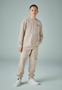 Beige krēmkrāsas džemperis un saskanīgas bikses, kas izgatavotas no mīksta auduma, ar taisnstūra formu un logotipa akcentiem. Apģērbu noslēdz sporta apavi.