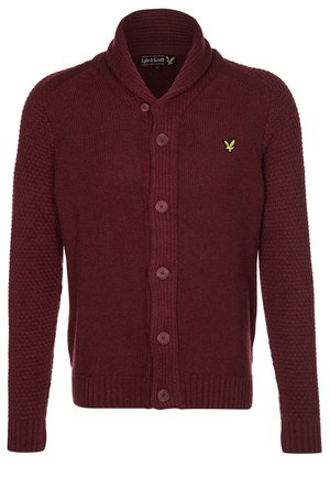 Man wearing black hooded jacket over burgundy sweater, dark jeans with cuffs, brown lace-up boots, and brown leather backpack straps.