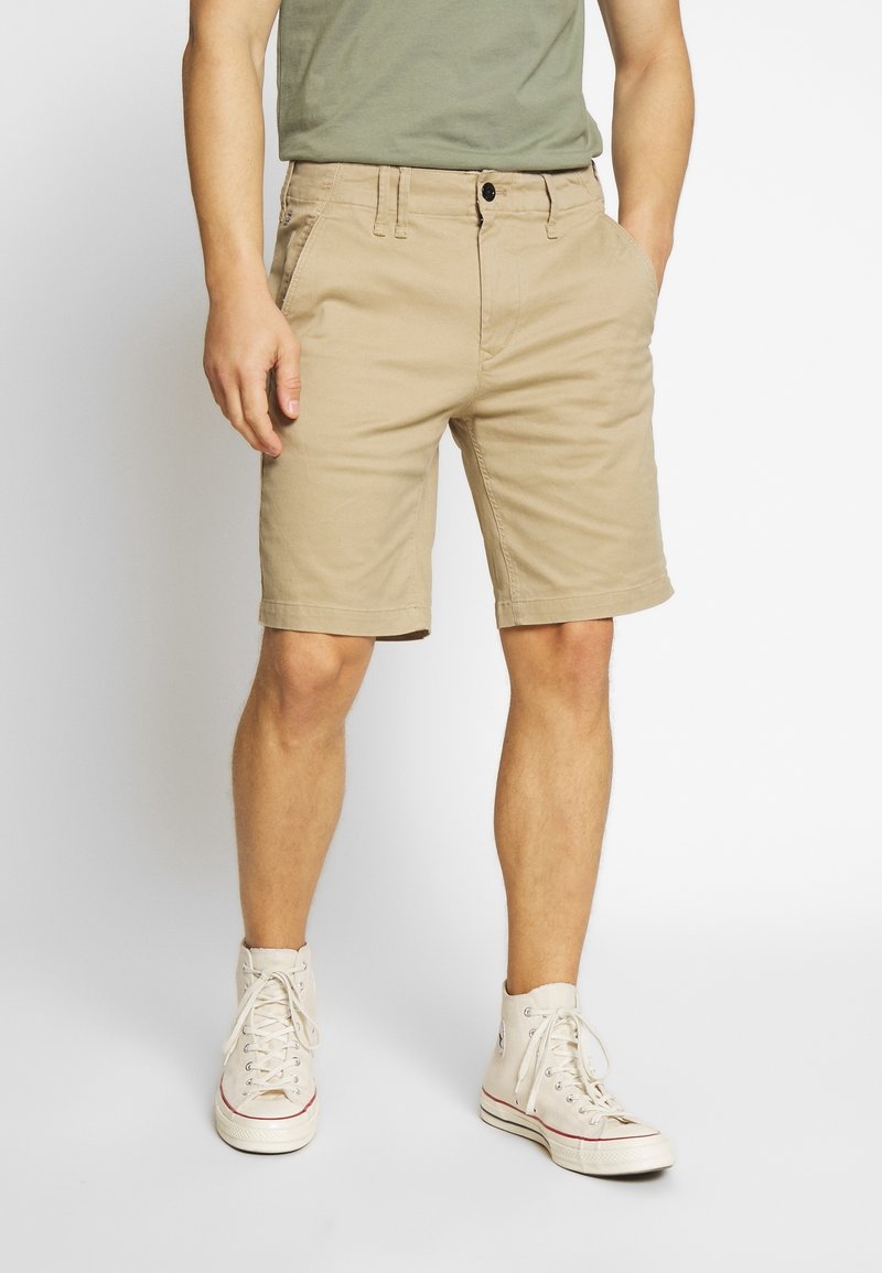 Pantaloni corti in cotone beige con una vestibilità rilassata, passanti per cintura, tasche frontali e chiusura con bottone. Indossati con sneakers bianche alte.