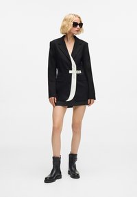 Melna žakete ar baltu centrālo paneļa daļu, ar vienu pogu un jostu. Uzvilkta virs īsas svārku kleitas un kombinēta ar melnām potīšu zābakām.