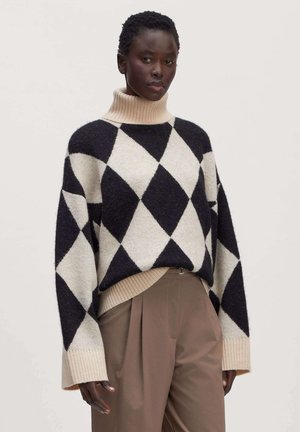 Vrouw draagt een zwarte en beige coltrui met argyle-patroon en hoge bruine broek, staand tegen een effen achtergrond.