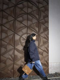 Navy puffer jacket, blue denim jeans, and brown suede handbag against a textured, geometric brown wall with triangular patterns.