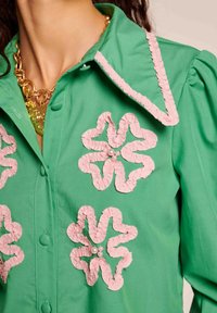 Zelena srajca s preklopom, ki vsebuje velike roza cvetje z nabranimi aplikacijami in bisernimi poudarki, ter ovratnik s podobnim roza obrobom.