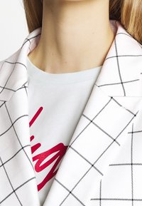 Blazer à carreaux blancs avec col superposé sur un t-shirt léger bleu clair à col rond présentant un grand motif graphique rouge. Tissu lisse et léger.