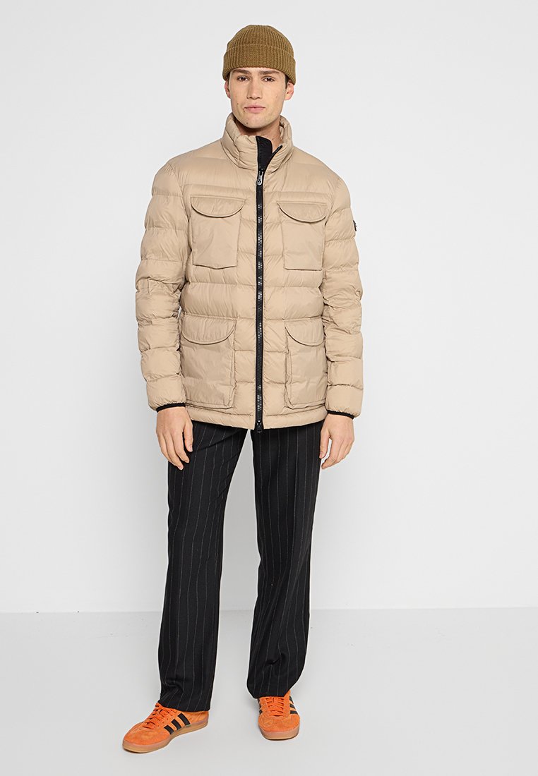 Beige puffjacka med hög krage, dragkedjestängning och fyra framfickor. Bärs med svarta rutiga byxor och orangea sneakers. Stickad brun mössa.