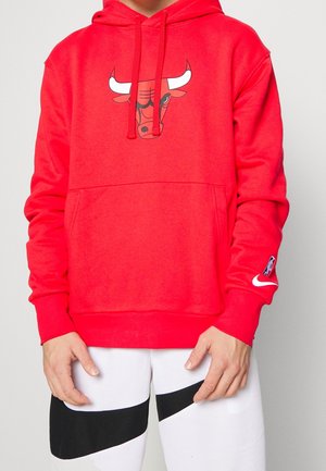 Personne portant un sweat à capuche rouge avec le logo des Chicago Bulls sur la poitrine, les logos Nike et NBA sur la manche, et un pantalon blanc avec des rayures diagonales noires.