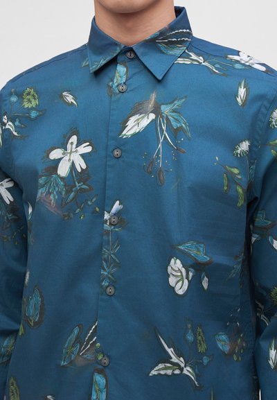 Άνδρας που φοράει μπλε πουκάμισο με κουμπιά και λευκό και πράσινο λουλουδάτο σχέδιο, στέκεται μπροστά σε ένα απλό φωτεινό φόντο.
