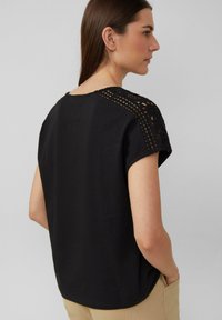 Haut noir à manches courtes avec un détail en crochet sur les épaules, coupe décontractée et texture lisse. Vue de dos présentée.