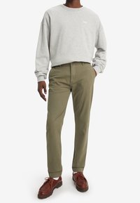 Grå sweatshirt med vit logotyp, olivgröna byxor och bruna snörskor, står på en enkel bakgrund med en avslappnad hållning.