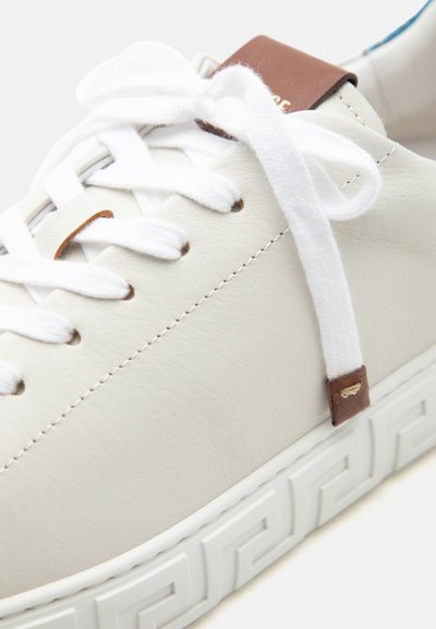 Gros plan d'une chaussure de sport en cuir blanc avec des lacets blancs, des accents en cuir marron et une semelle blanche présentant un motif géométrique.