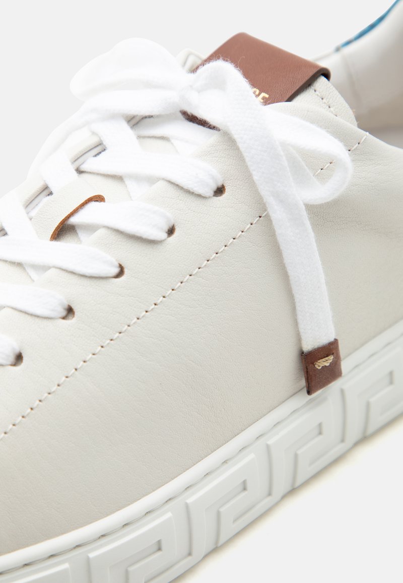 Gros plan d'une chaussure de sport en cuir blanc avec des lacets blancs, des accents en cuir marron et une semelle blanche présentant un motif géométrique.