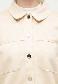 Camicia color crema con colletto arrotondato, grandi tasche con patta e quattro lucidi bottoni metallici sulla parte anteriore.