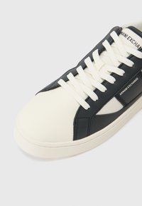 Hvit og marineblå lavtopp sneakers med en glatt læroverdel, kontrasterende teksturer, signaturmerke og hvite lisser. Flat gummisåle.