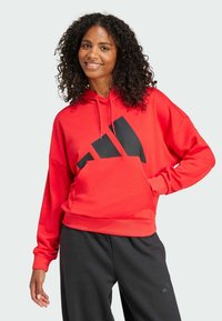 Röd hoodie med svart grafisk design som har tre ränder. Tillverkad av ett mjukt material, har den en avslappnad passform och framficka.
