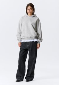 Szara bluza z kapturem z przednią kieszenią, noszona na białej koszulce; połączona z luźnymi, ciemnymi dżinsami z kontrastowymi przeszyciami.