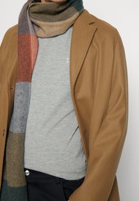 Cappotto di lana tan sopra una maglietta grigia, completato da un sciarpa a quadri multicolore con sfumature di verde, arancione e marrone.