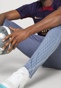 Conjunto deportivo que incluye una camiseta de color púrpura oscuro con detalles rojos, leggings azules con estampado y zapatillas blancas con detalles en turquesa, sosteniendo un balón de fútbol.