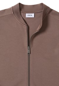 Veste taupe à fermeture éclair argentée partiellement ouverte, révélant une étiquette blanche avec le texte "CECIL" à l'intérieur du col.