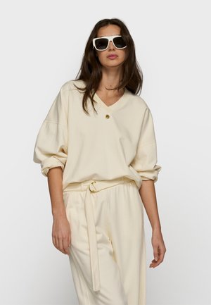 Vrouw draagt een oversized crèmekleurige trui en bijpassende broek met hoge taille en riem, geaccentueerd met grote witgerande zonnebril.