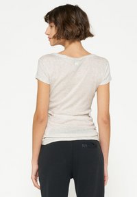 Lichtgrijze, aansluitende t-shirt met korte mouwen, zachte textuur, brede halslijn en subtiele details op de rug, gedragen met zwarte broek.