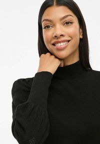 Jeune femme avec de longs cheveux noirs portant un pull noir à col montant avec des manches bouffantes texturées, souriant avec la main reposant près de son menton.
